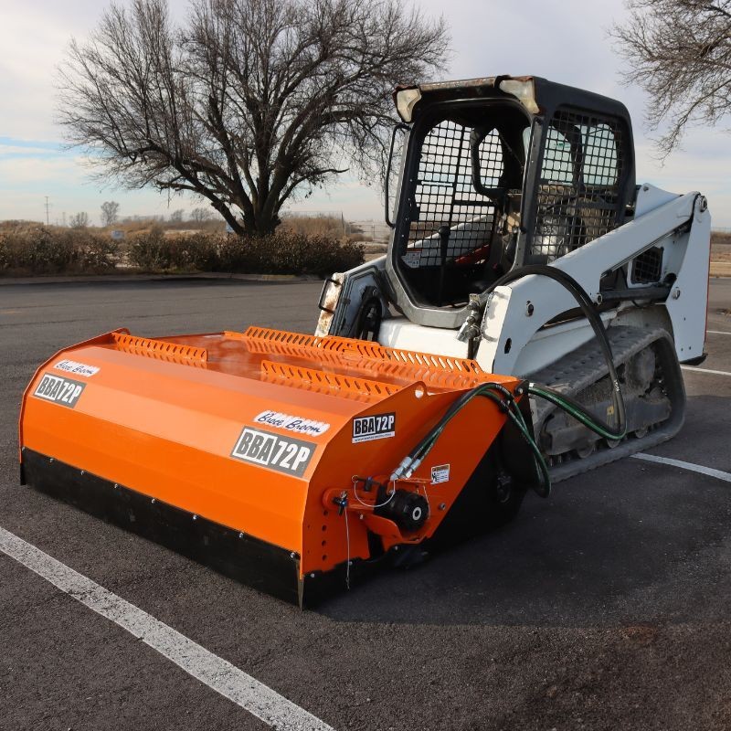 Bobcat loader with an orange attachment on an empty parking lot, trees in the background.